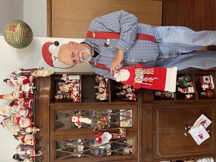 White man with white goatee stands in front of shelving with large number of Santa figurines.