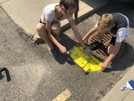 Two young people painting a yellow fish stencil onto the road near a storm drain